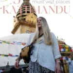 Grafika na Pinterest (Pin) Kobieta z długimi blond włosami i czerwonym tiką na czole stoi przed stupą Swayambhunath (Monkey Temple) w Katmandu, Nepal. W tle widoczne są kolorowe flagi modlitewne, złota wieża świątyni oraz lokalni mieszkańcy i stragany z pamiątkami.