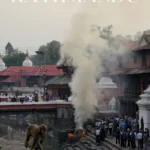 Grafika Pinterest (Pin) Widok na kremację nad rzeką Bagmati w świątyni Pashupatinath w Katmandu, Nepal. Dym unosi się nad płonącym stosie pogrzebowym, wokół którego zgromadzeni są ludzie. Na pierwszym planie widać małpę spacerującą po murku.