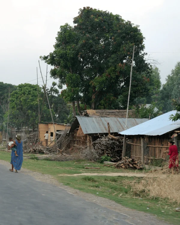 Tharu – rdzenna ludność Chitwan w Dolinie Teraj, znani jako ludzie lasu. Kobieta w tradycyjnym stroju niesie dziecko obok chaty krytej blachą, w tle wiejska zabudowa i drzewa.